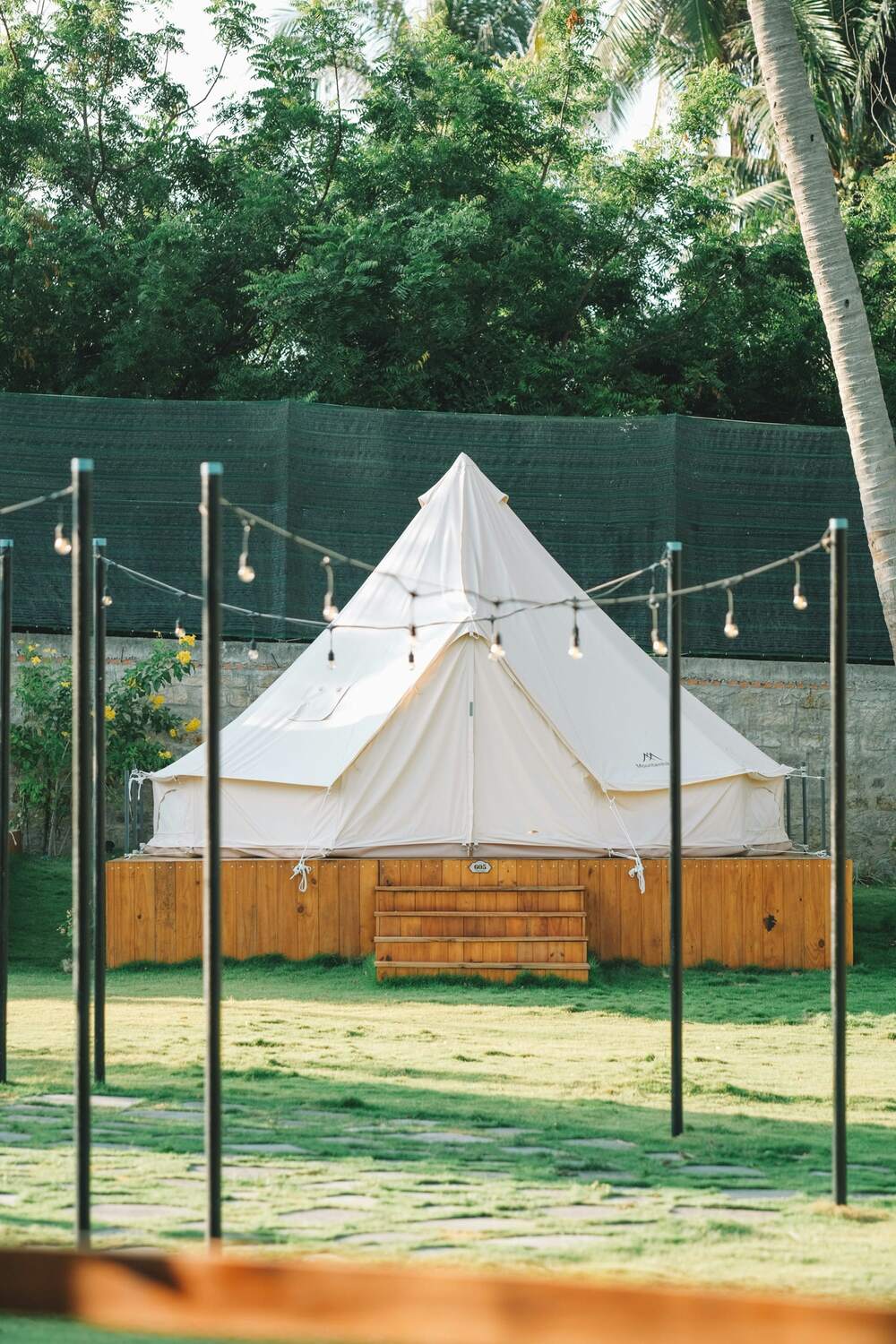 A canvas bell tent on a wooden deck, strung with festoon lights among palm trees